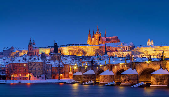 Central European Ring Prague Castle illuminated at twilight with snow-covered roofs and Charles Bridge spanning the frozen Vltava River